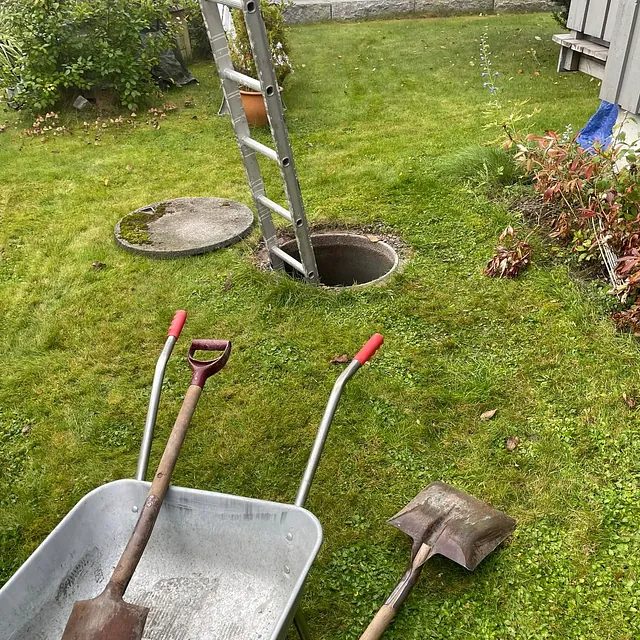 En hage med en betongbrønn og en metallstige som fører ned i den, omgitt av en trillebår og spader på gresset.