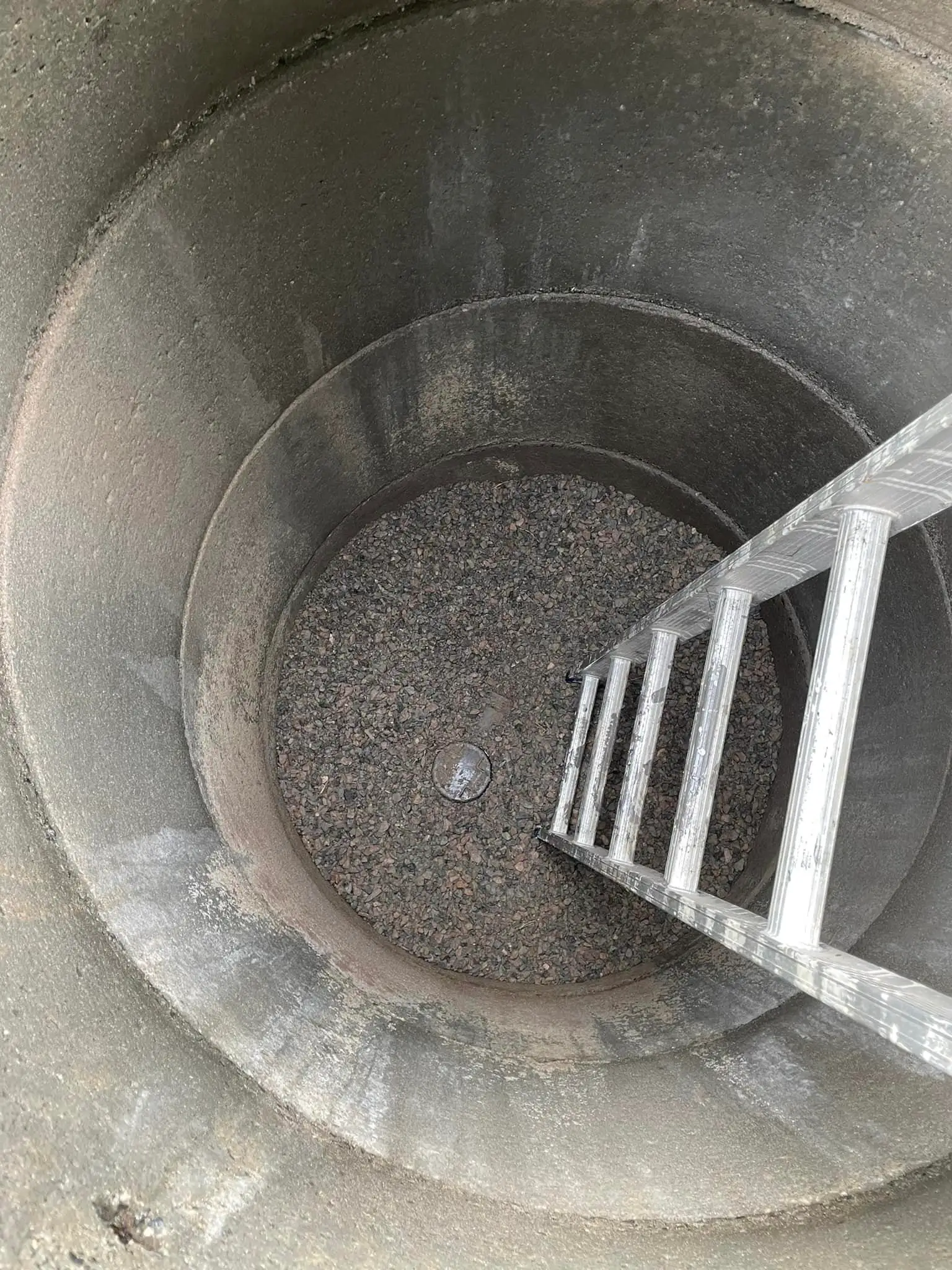 En betongbrønn med en stige som strekker seg ned til bunnen, dekket av grus.