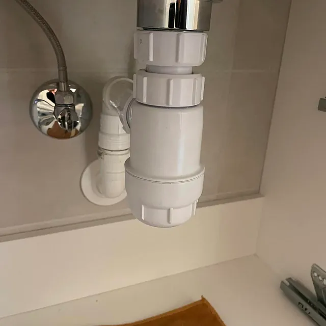 Nærbilde av et hvitt avløpsrør under en vask, med en krom kran på veggen i bakgrunnen.