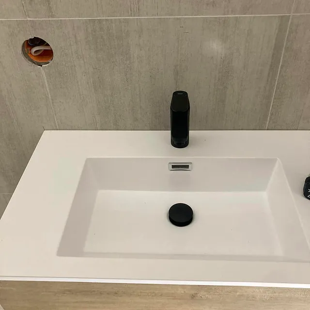 En hvit rektangulær servant med en svart kran og et sort avløpslokk, montert mot en lys flislagt vegg med et åpent hull.