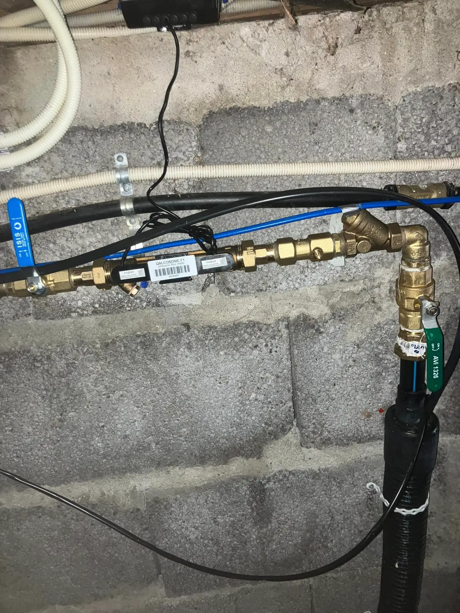 Vannrør og koblinger montert på en vegg av ubehandlede murblokker, med elektriske ledninger og klemmer.
