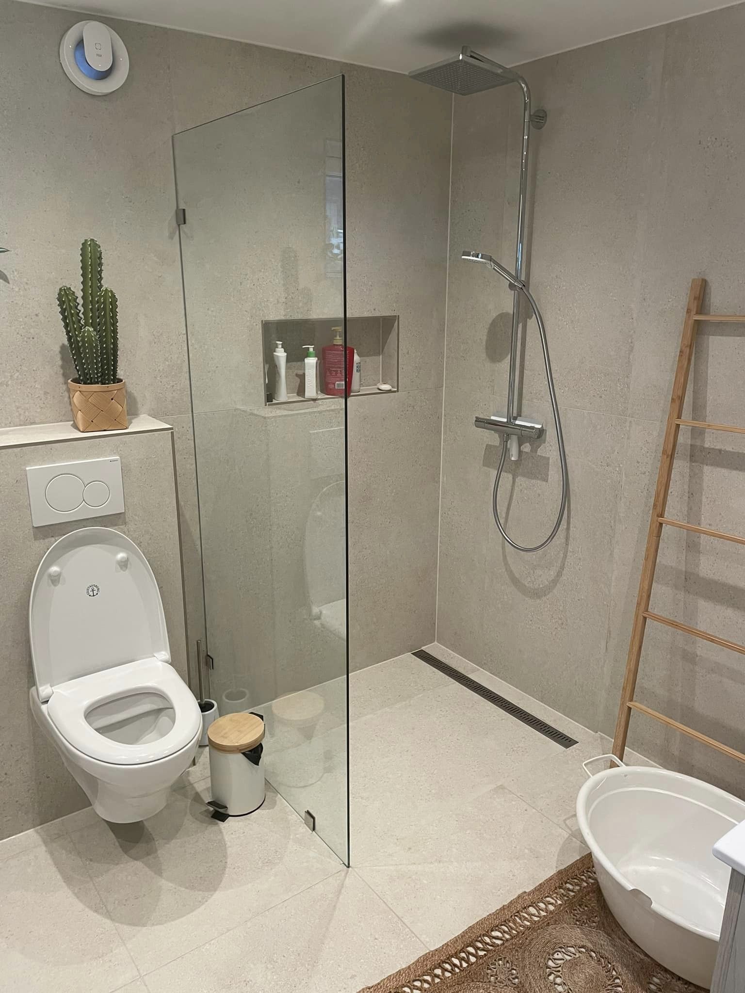 Bad med dusjkabinett av glass, vegghengt toalett, og en kaktus på en hylle, med lys grå fliser på gulvet og veggene.