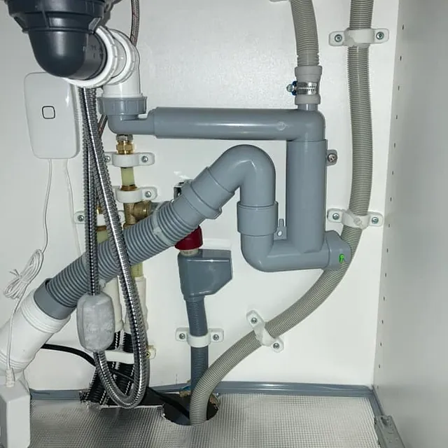 Rørinstallasjoner under en vask med flere sammenkoblinger av grå rør og slanger.